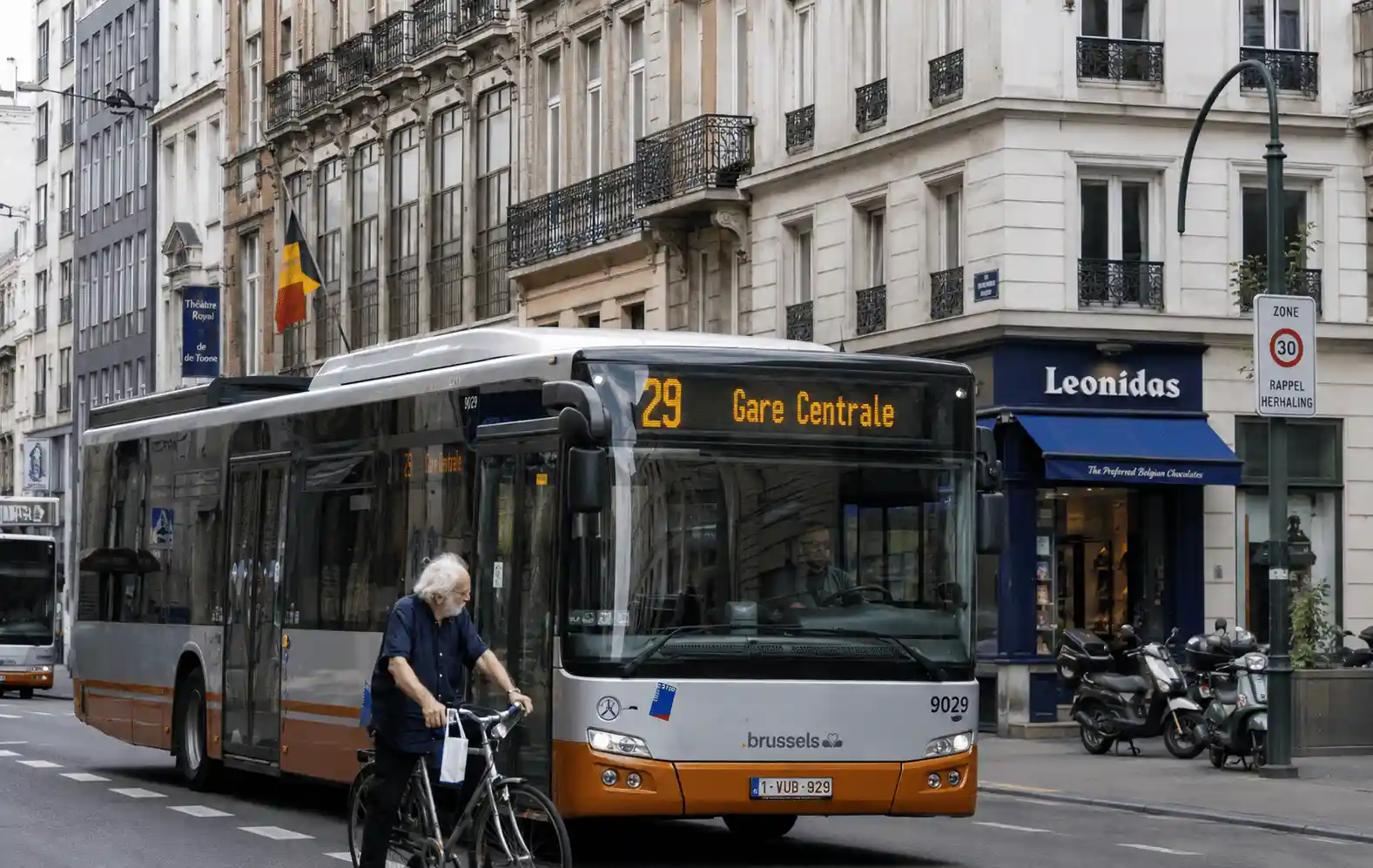Un cycliste circule tout près de l’avant d’un bus dans une rue urbaine ; un panneau rappelle la zone 30.