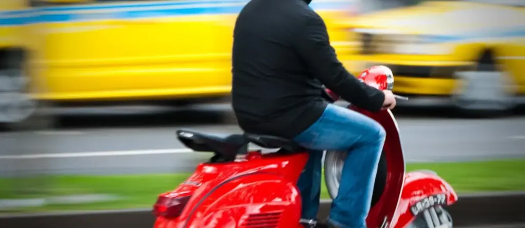 Conducteur sur un scooter rouge, avec un bus jaune en arrière-plan.