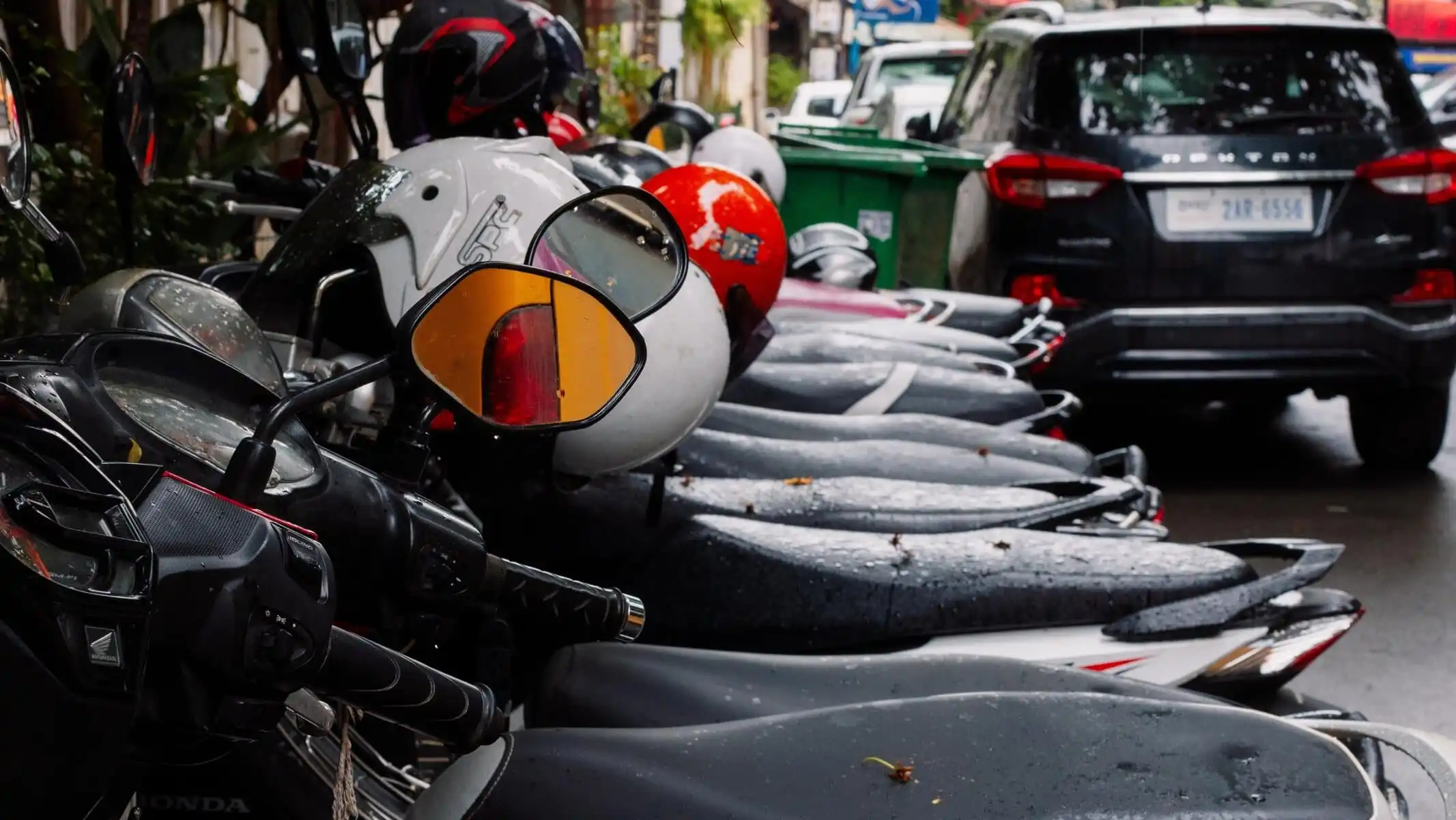 Scooters stationnés en rang, aux selles couvertes de gouttes d’eau, au bord d’une chaussée mouillée.