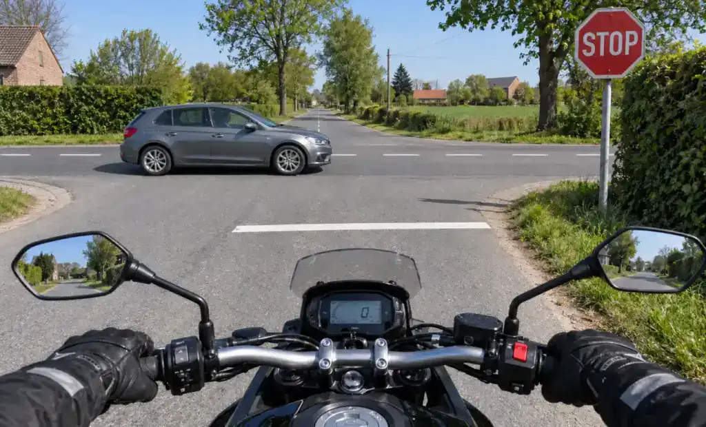 Vue depuis une moto arrêtée avant la ligne d’arrêt à un STOP, tandis qu’une voiture traverse l’intersection de gauche à droite.