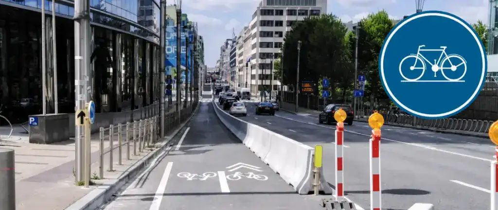 Piste cyclable séparée de la chaussée par des barrières en béton, avec vélos et chevrons peints au sol et panneau rond bleu à vélo blanc en incrustation.