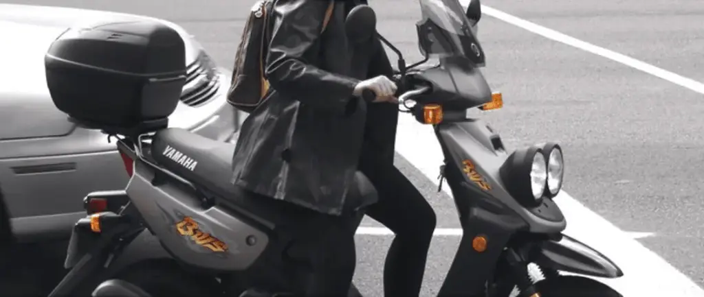 Conducteur sur un scooter gris équipé d’un coffre arrière, à côté d’une voiture.