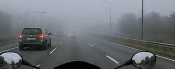 Vue depuis une moto sur une autoroute dans le brouillard, avec une voiture devant à gauche et d’autres véhicules à peine visibles au loin.