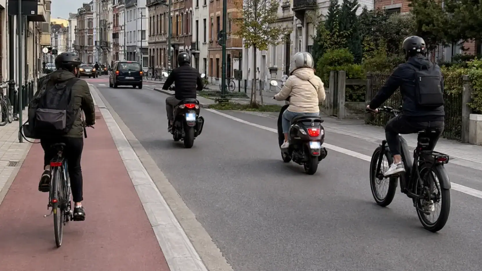 Dans une rue, un cycliste roule sur une bande rouge à gauche, tandis que deux scooters et un autre cycliste circulent sur la chaussée.