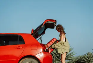 Une personne manipule une valise près du coffre ouvert d’une voiture.