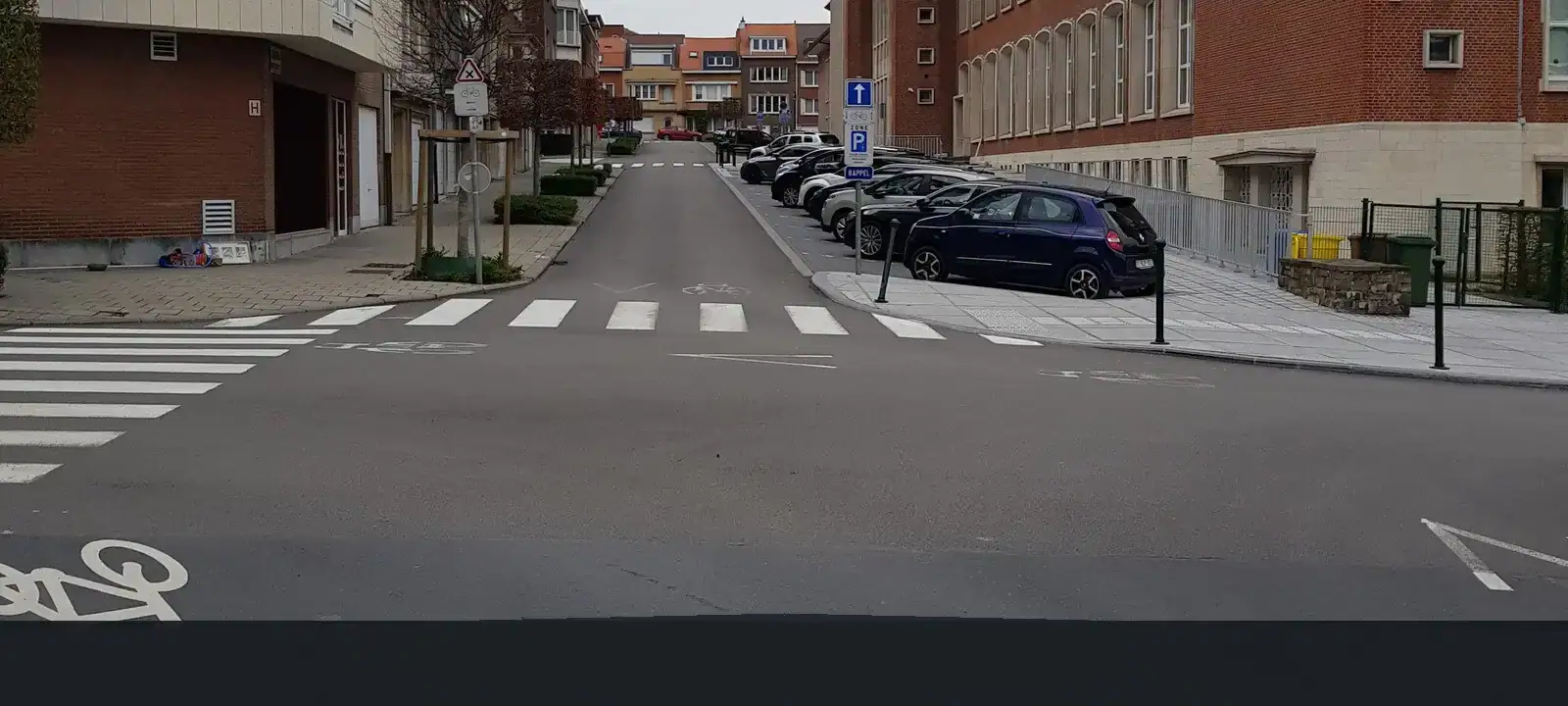 Carrefour entre des bâtiments proches de la chaussée, avec des passages pour piétons et des voitures stationnées dans la rue en face.