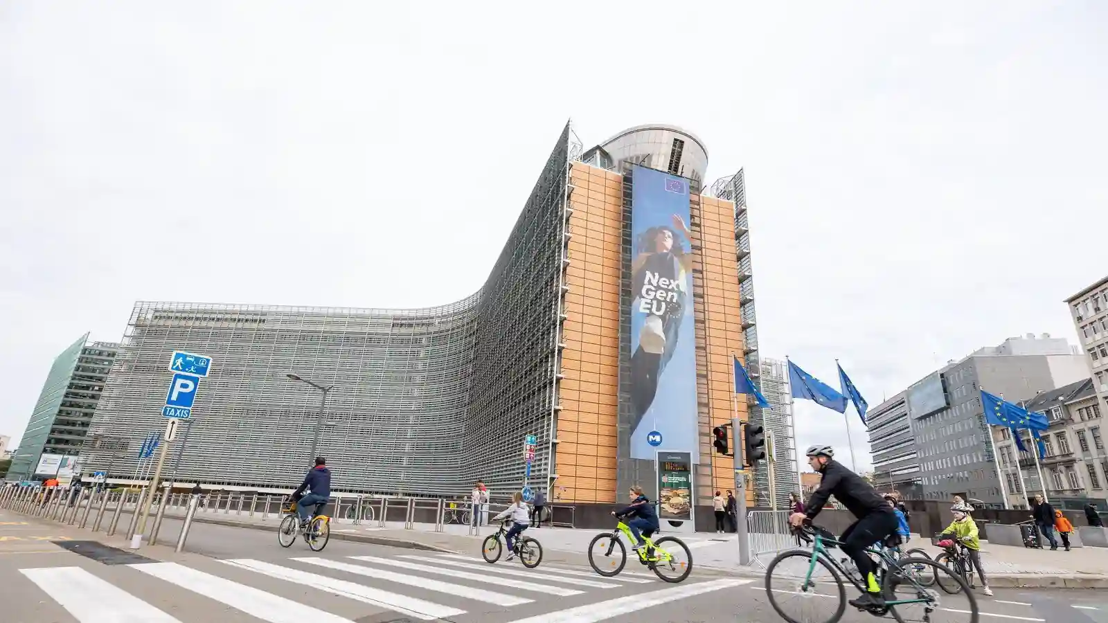 Un cycliste au premier plan à droite, des enfants à vélo au milieu et d’autres cyclistes plus loin à gauche, près d’un passage pour piétons ; des piétons se tiennent sur le trottoir.
