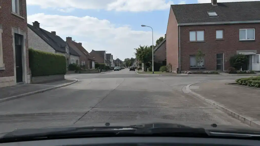 Vue depuis le poste de conduite vers un carrefour entre des rues bordées de maisons, sans agent, feu ni signal de priorité visible.