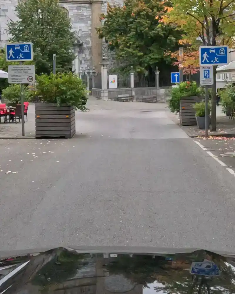 Vue depuis une voiture vers une place dont l’entrée est signalée par deux panneaux de zone résidentielle ou de rencontre.