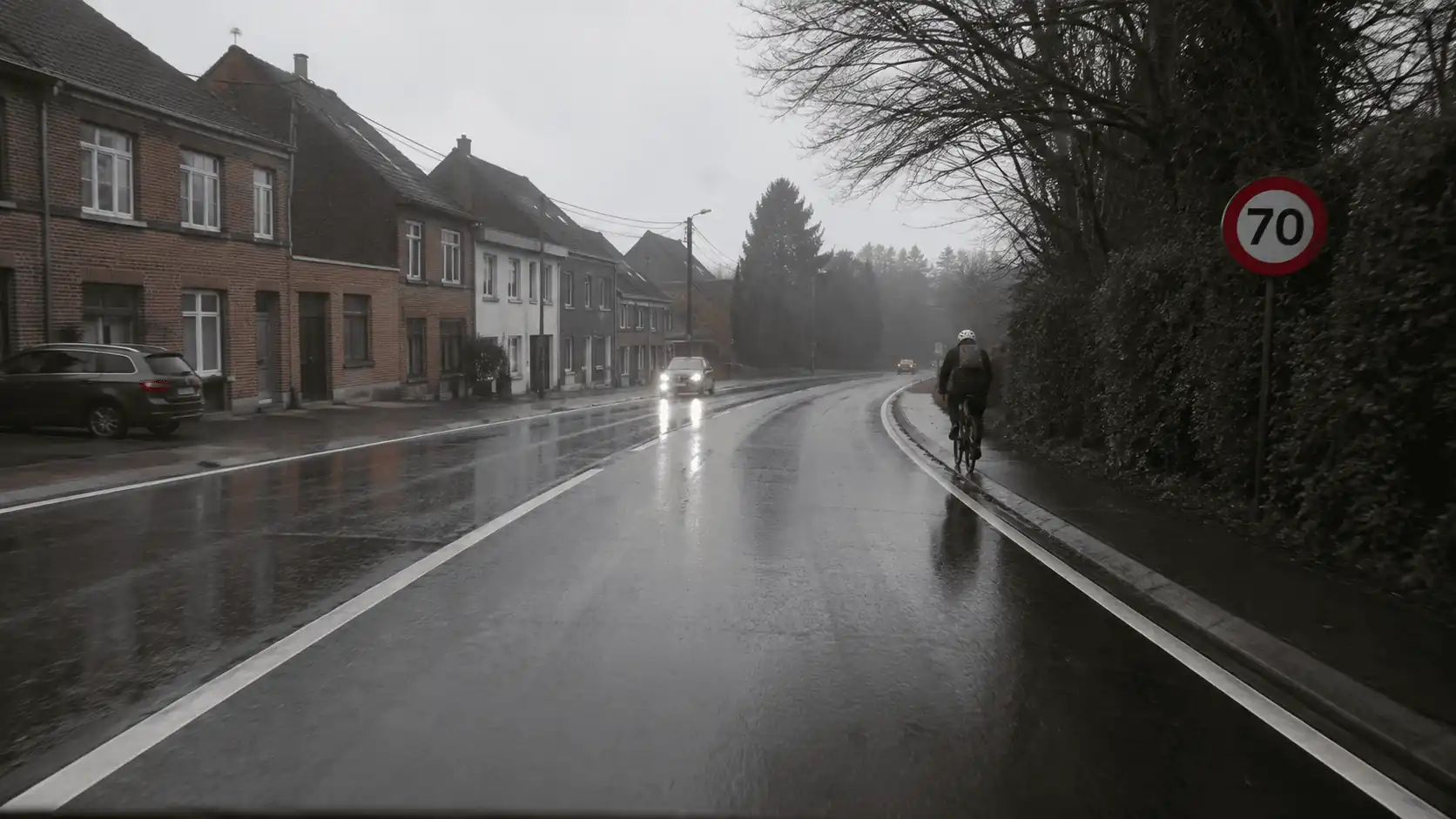 Chaussée mouillée dans un virage, avec un cycliste au bord droit, une voiture en sens inverse et un panneau de limitation à 70 km/h.