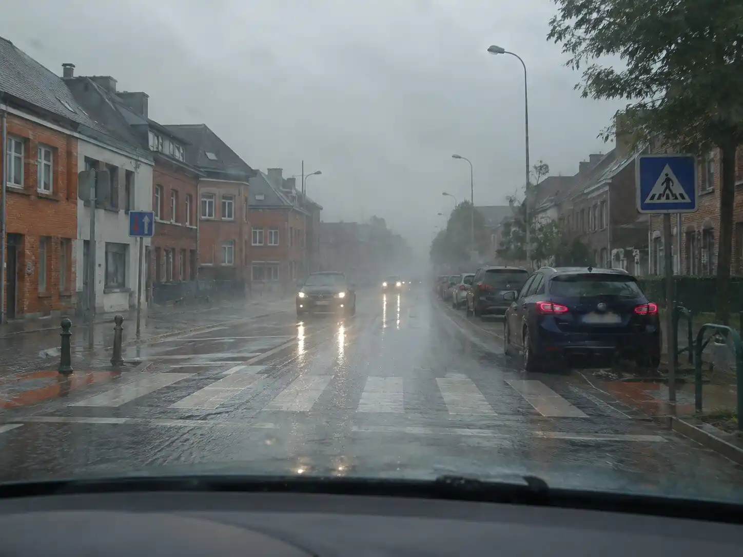 Rue sous une forte pluie, avec un passage pour piétons, des reflets de phares sur la chaussée mouillée et une visibilité réduite au loin.