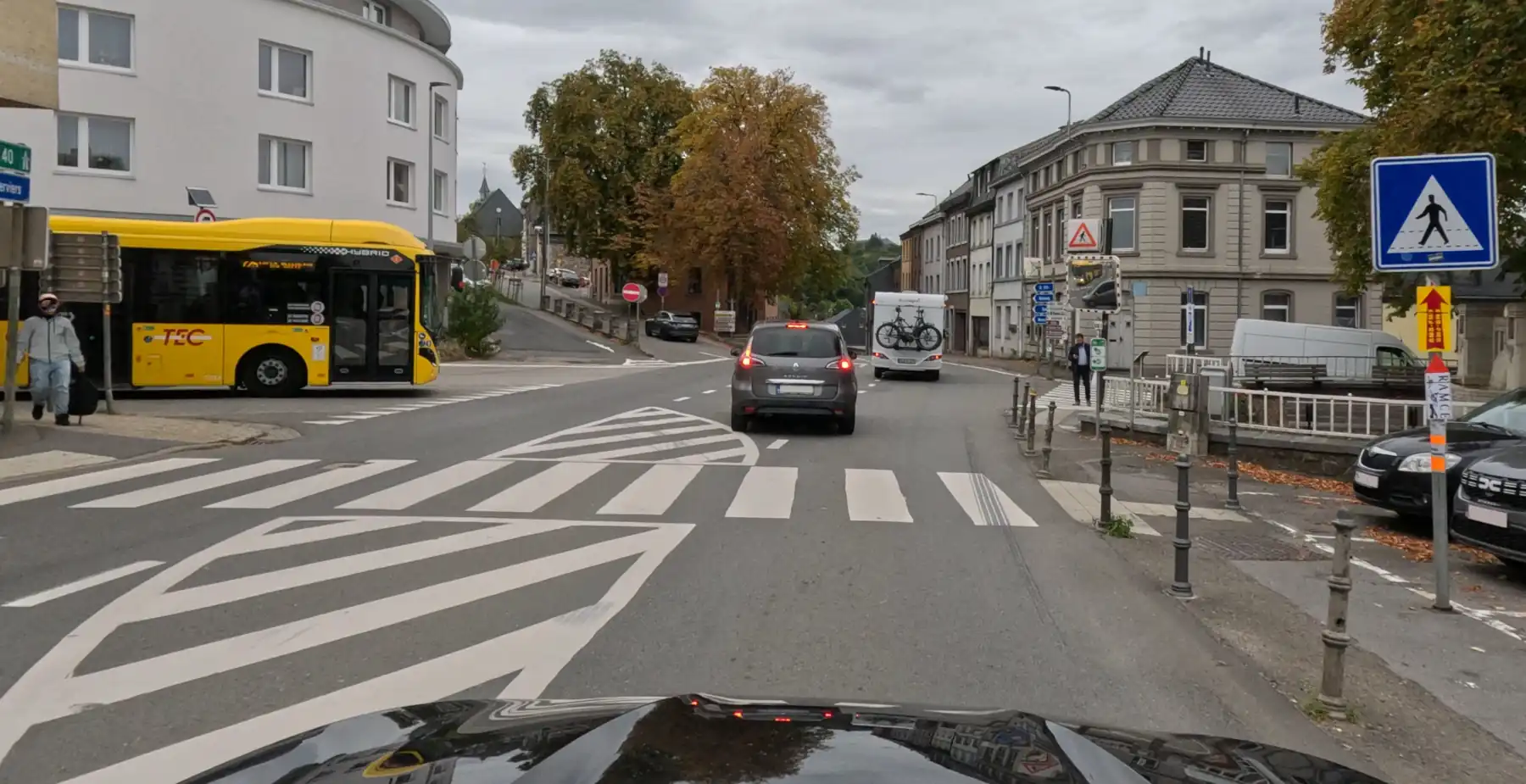 Carrefour urbain avec une voiture à droite d’une zone hachurée, un passage pour piétons et un bus à gauche.