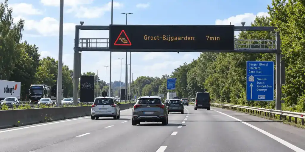 Plusieurs véhicules sur trois bandes, sous un panneau lumineux affichant un pictogramme de file de véhicules dans un triangle rouge et « Groot-Bijgaarden: 7min ».