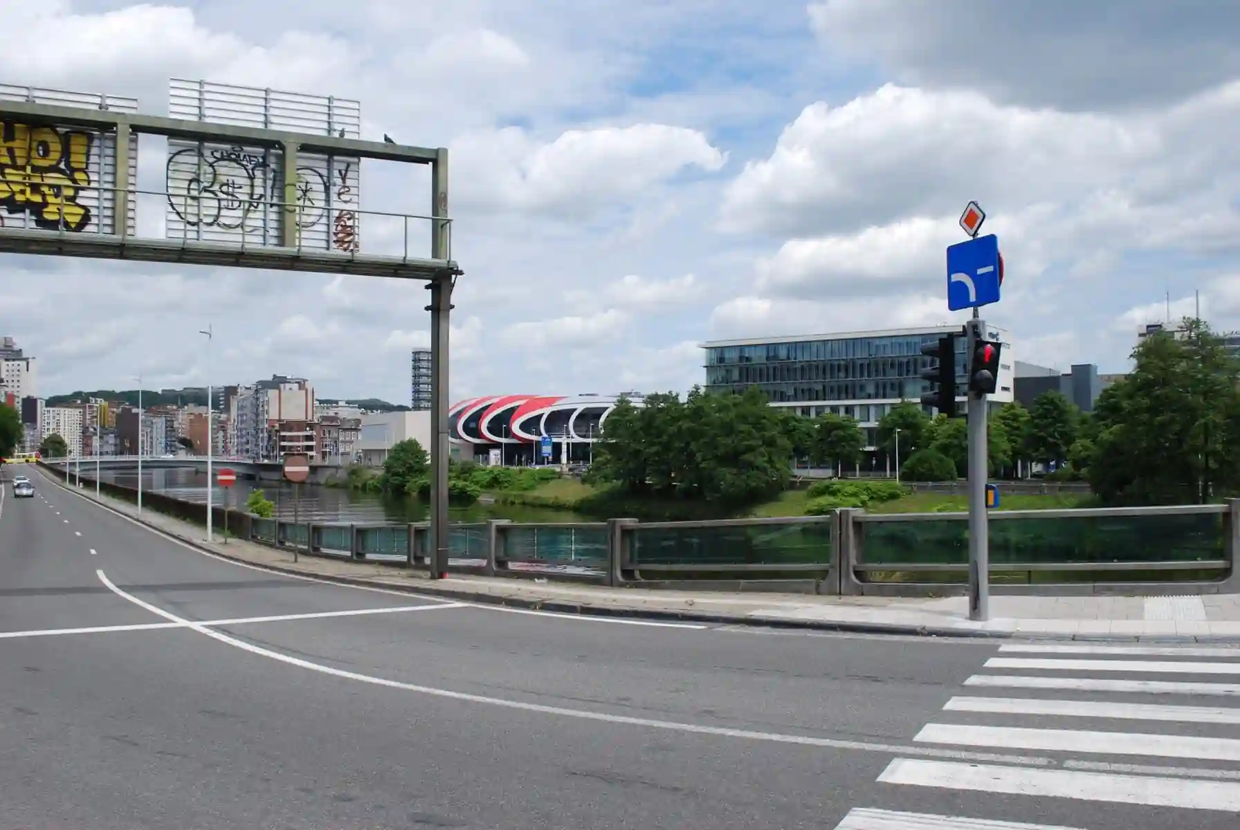 Signal à deux optiques avec silhouette rouge de piéton, sous un panneau en losange et un panneau bleu au tracé blanc coudé vers la gauche, au bord d’une chaussée courbe.