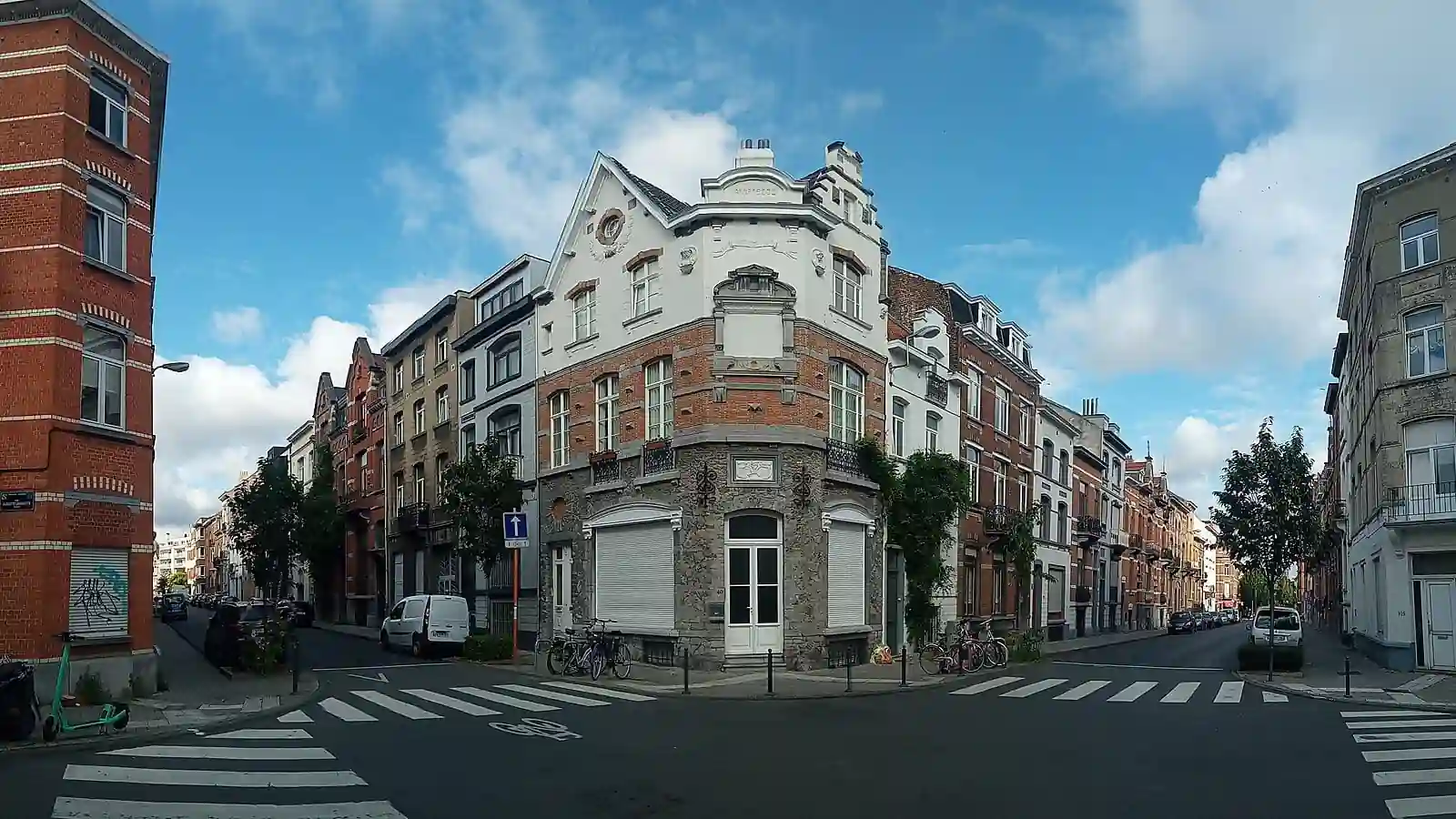 Carrefour avec un immeuble d’angle au centre, des passages pour piétons et une camionnette blanche stationnée près du trottoir dans la rue de gauche.