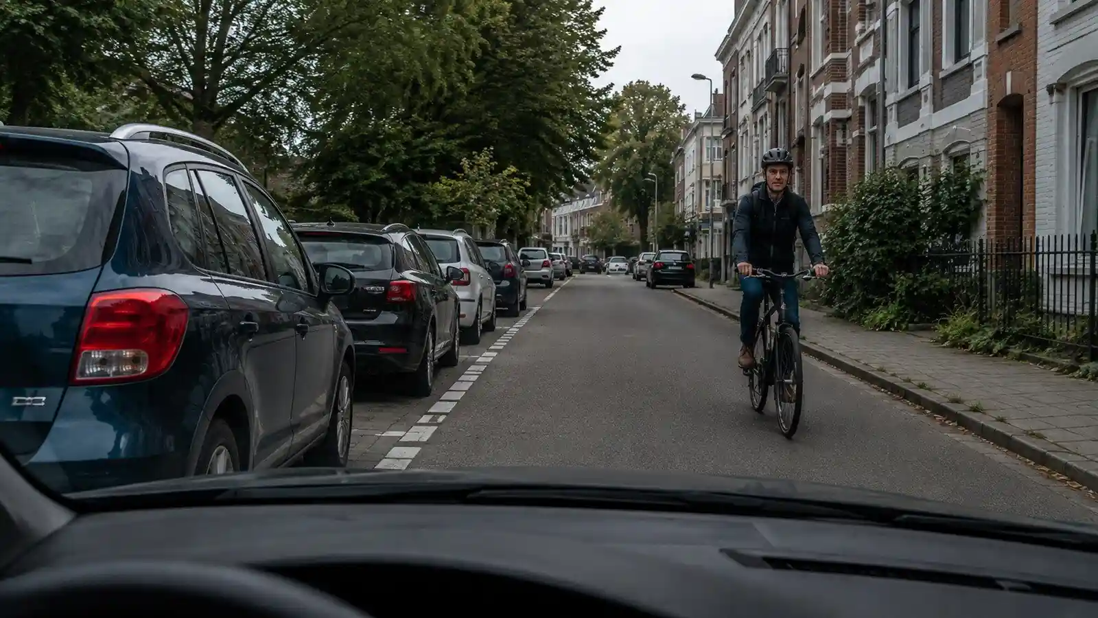 Un cycliste vient de face sur la droite de l’image ; une rangée de voitures stationnées borde le côté gauche de la chaussée.