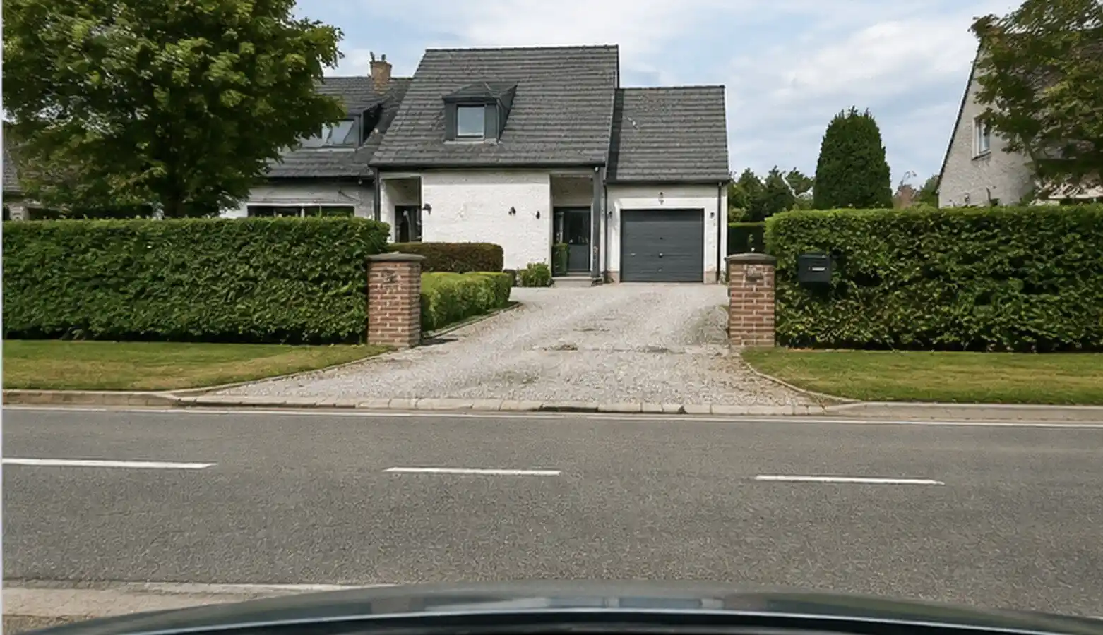 Entrée pavée d’une maison avec garage, entre deux piliers, donnant sur une chaussée à ligne axiale discontinue.
