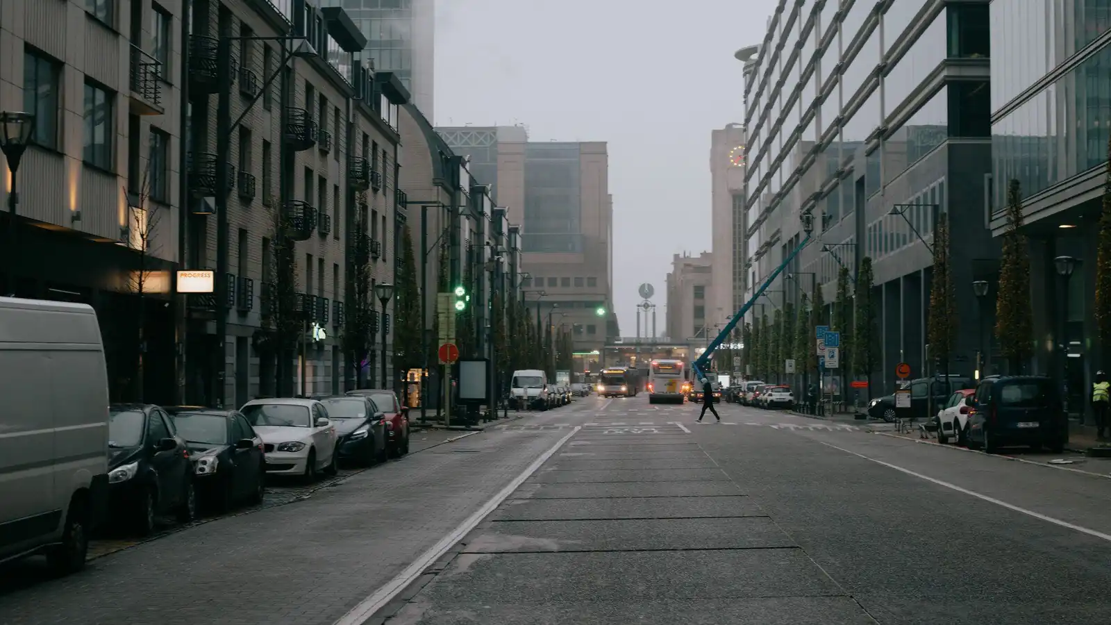 Rue brumeuse bordée de véhicules stationnés, avec un piéton traversant plus loin et des bâtiments dont les contours s’estompent avec la distance.