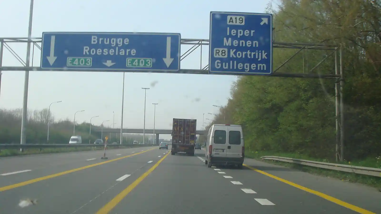 Des lignes jaunes continues délimitent un tracé décalé par rapport aux lignes blanches discontinues, avec des balises à gauche et un camion et une camionnette devant.