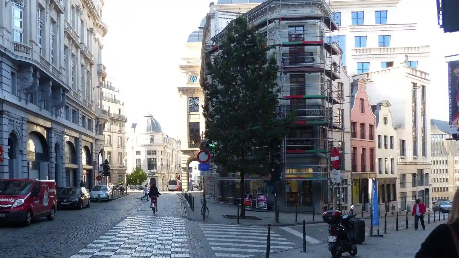 Rue pavée se divisant autour d’un bâtiment : un cycliste roule à gauche, un passage pour piétons traverse la branche de droite, avec des piétons et des véhicules à proximité.