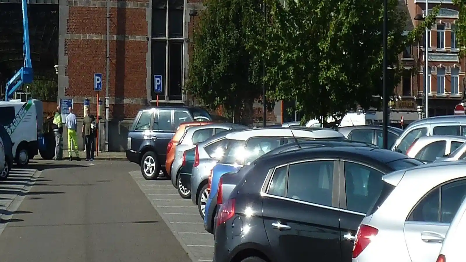 Allée de parking bordée de places marquées au sol et de voitures stationnées, avec des piétons à son extrémité.