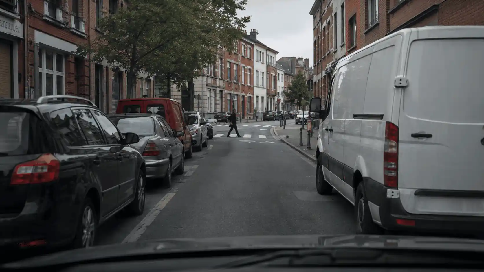 Rue bordée de véhicules stationnés, avec un grand utilitaire blanc masquant le côté droit et un piéton sur un passage plus loin.
