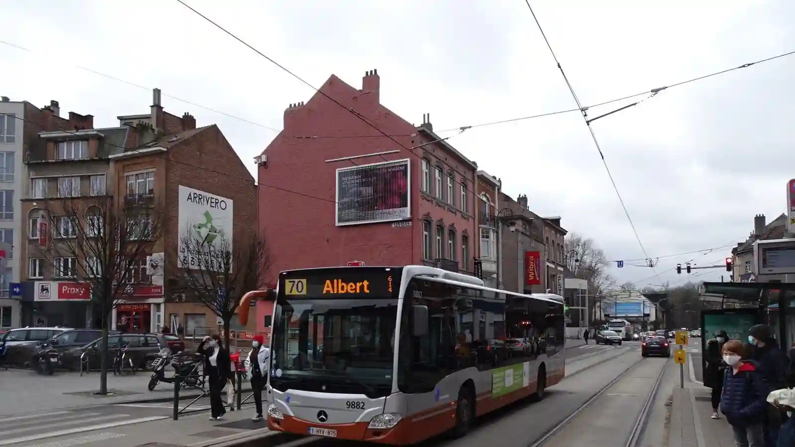 Un autobus occupe une grande partie du premier plan, près d’un passage pour piétons ; des personnes sont visibles près de l’avant du bus et sur le trottoir à droite.