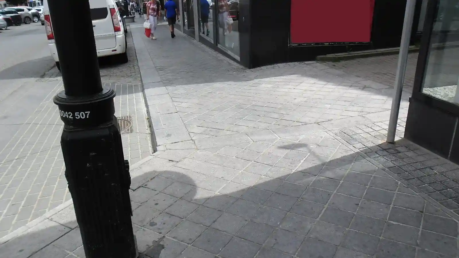 Trottoir pavé longeant des commerces, avec un lampadaire et des changements de revêtement.