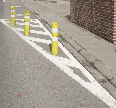 Zone délimitée par une ligne blanche continue et remplie de stries blanches obliques, avec trois potelets jaunes le long du trottoir.