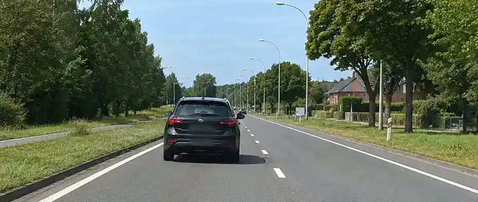 Route sèche avec une voiture devant, une vue dégagée au loin, des arbres sur les côtés et des maisons à droite.