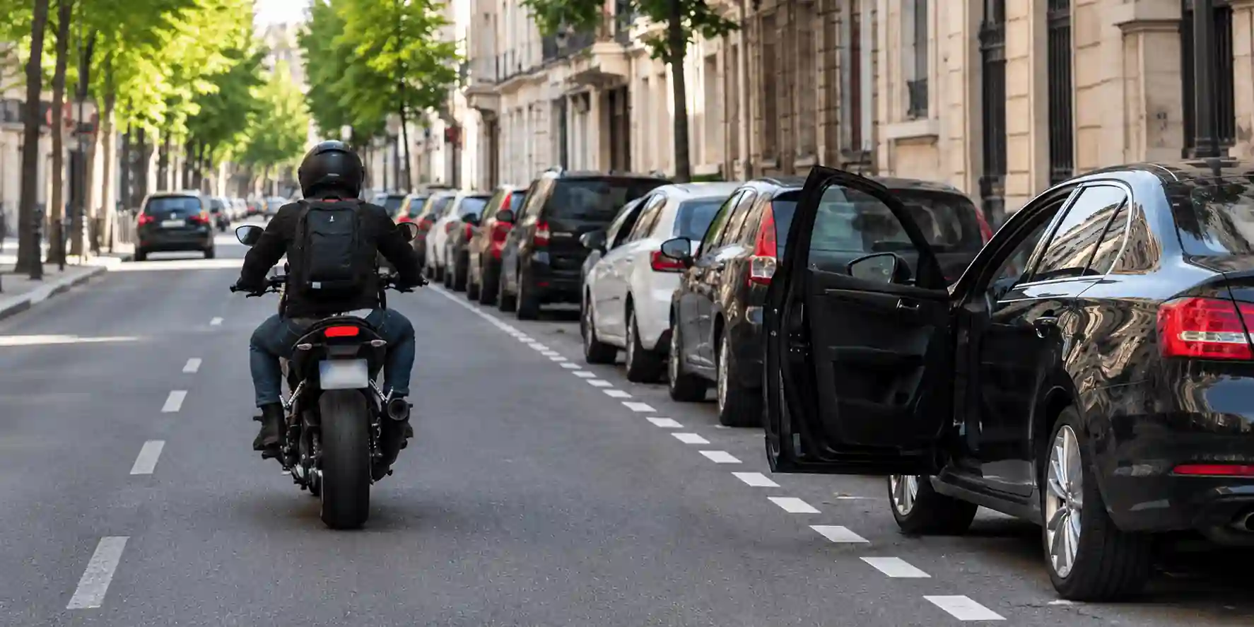 Un motocycliste longe des voitures stationnées à droite, dont l’une a la portière côté chaussée ouverte.