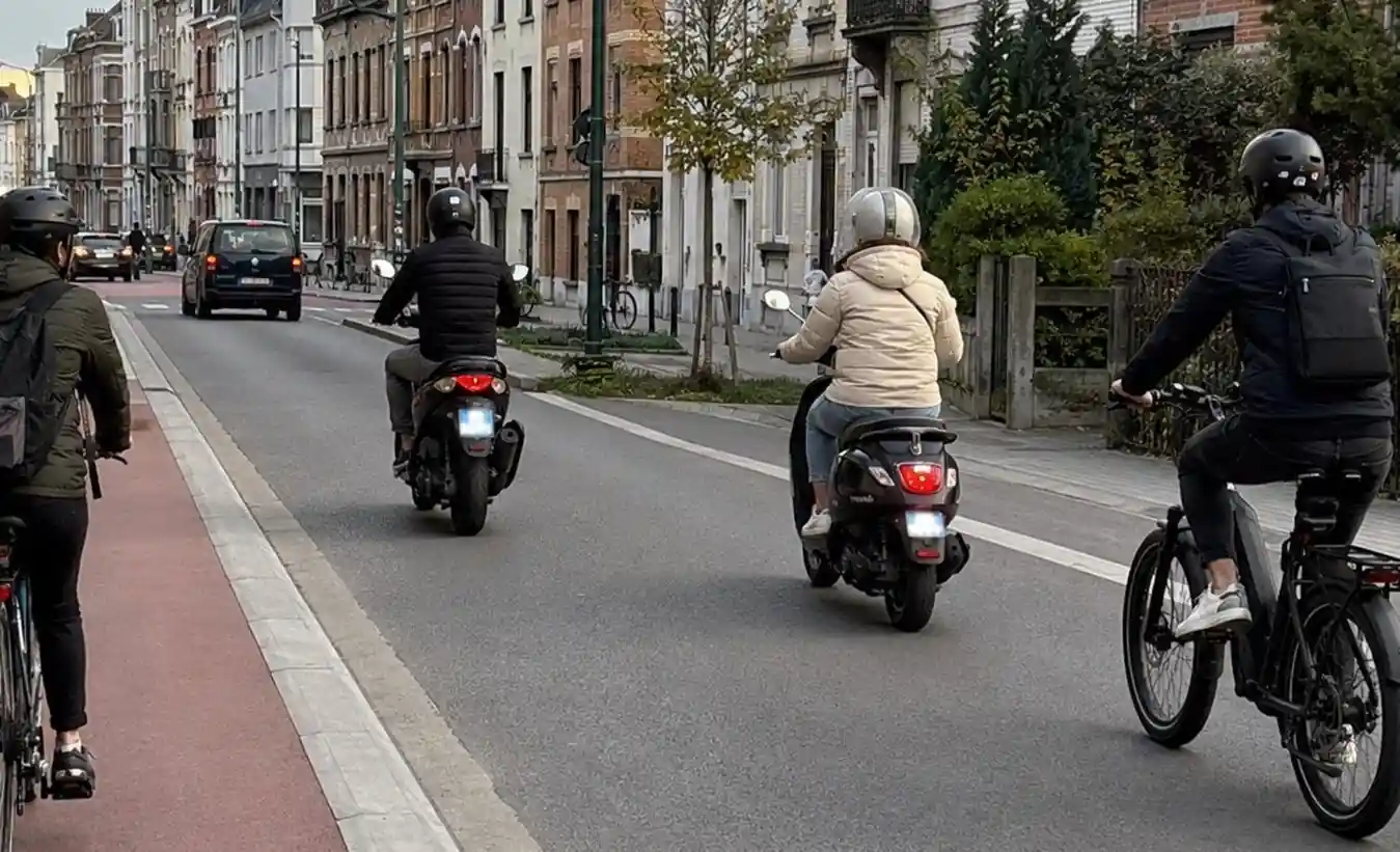 Deux scooters et un cycliste circulent sur la chaussée, avec des voitures devant ; un autre cycliste est visible sur une piste rouge à gauche.