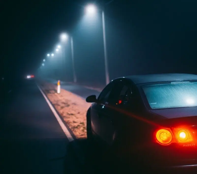 Voiture vue de l’arrière, feux arrière allumés, sur une route de nuit éclairée par des lampadaires.