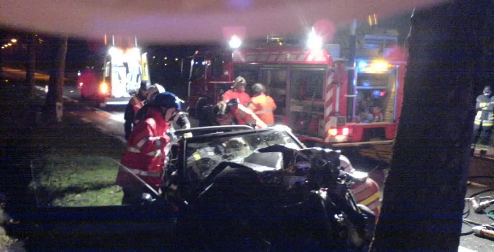 Secouristes autour d’une voiture très endommagée, de nuit, avec une ambulance et un camion de pompiers à proximité.