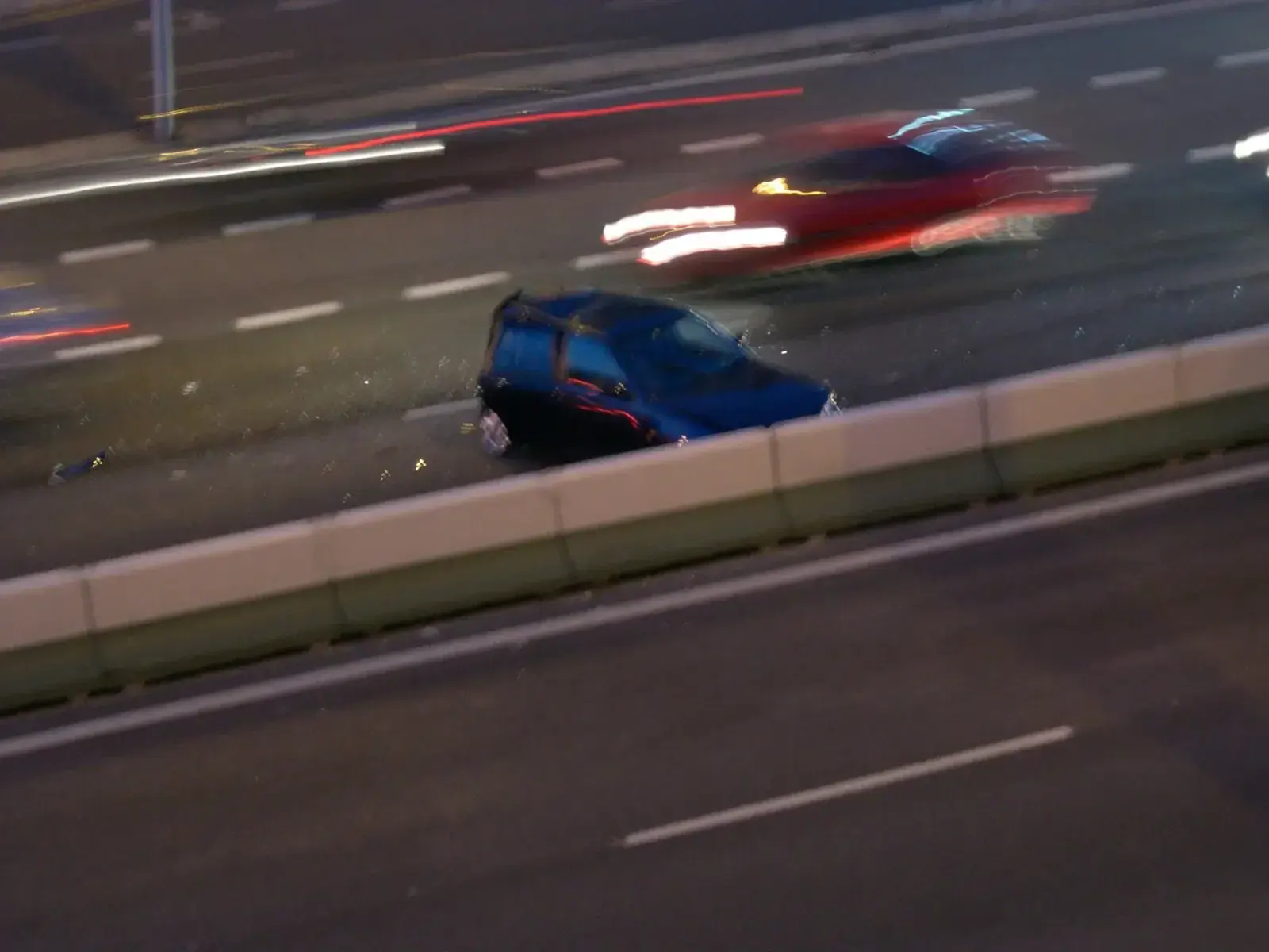 Voiture endommagée près d’une barrière en béton, de nuit, tandis que d’autres véhicules passent à proximité.