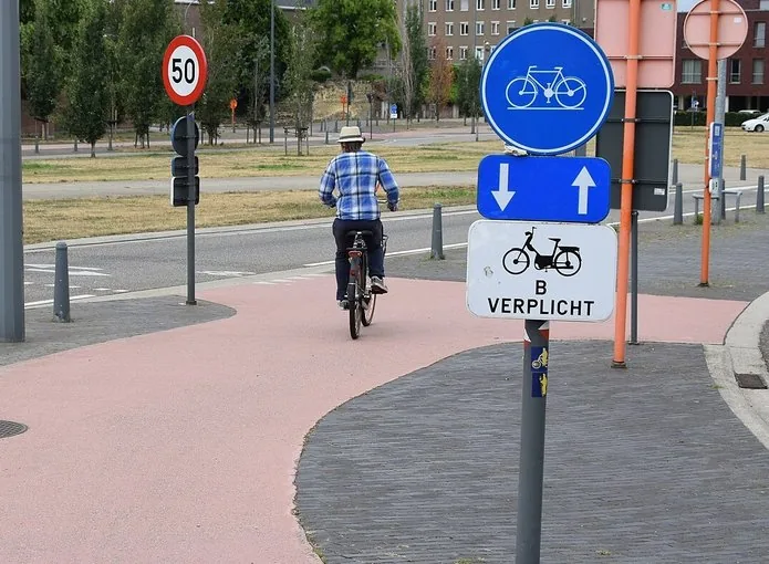 Cycliste sur une piste signalée par un disque bleu avec un vélo blanc. En dessous figurent deux flèches opposées et un panneau portant un symbole de cyclomoteur et « B VERPLICHT ».