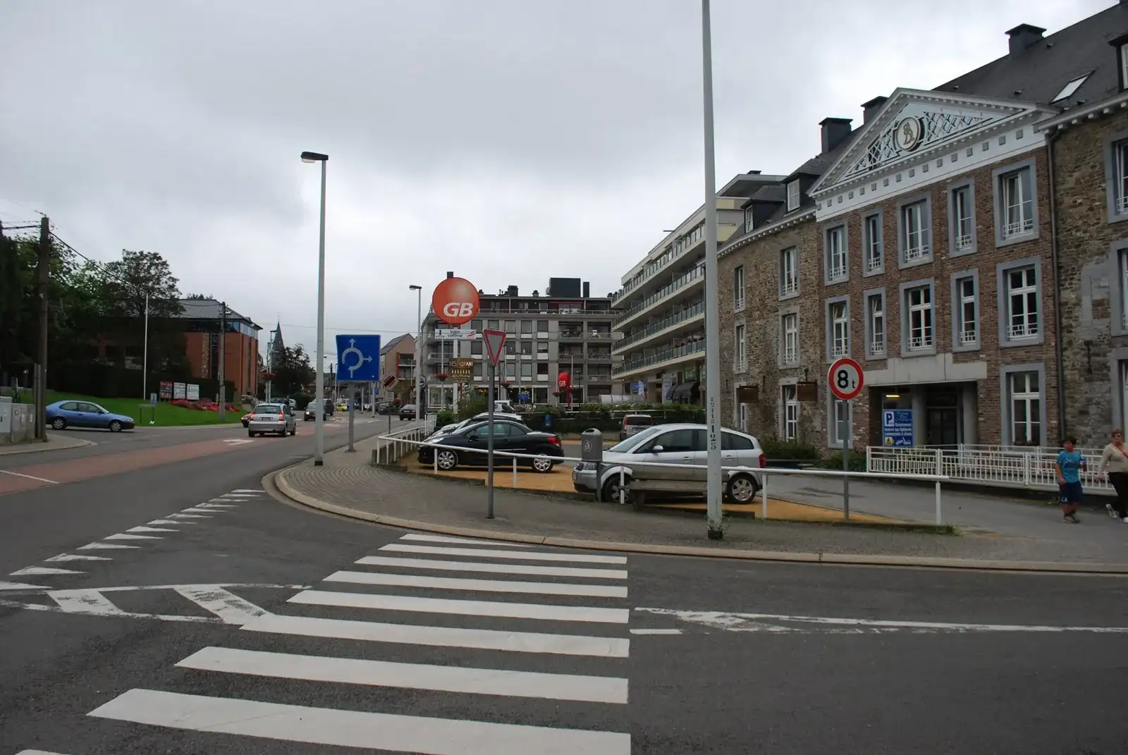 Carrefour à Embourg avec passage piéton, signal B1 et triangles au sol, préavis de rond-point F25, fin de voie prioritaire B11 et panneau « 8 t ». Des personnes se trouvent sur le trottoir à droite.