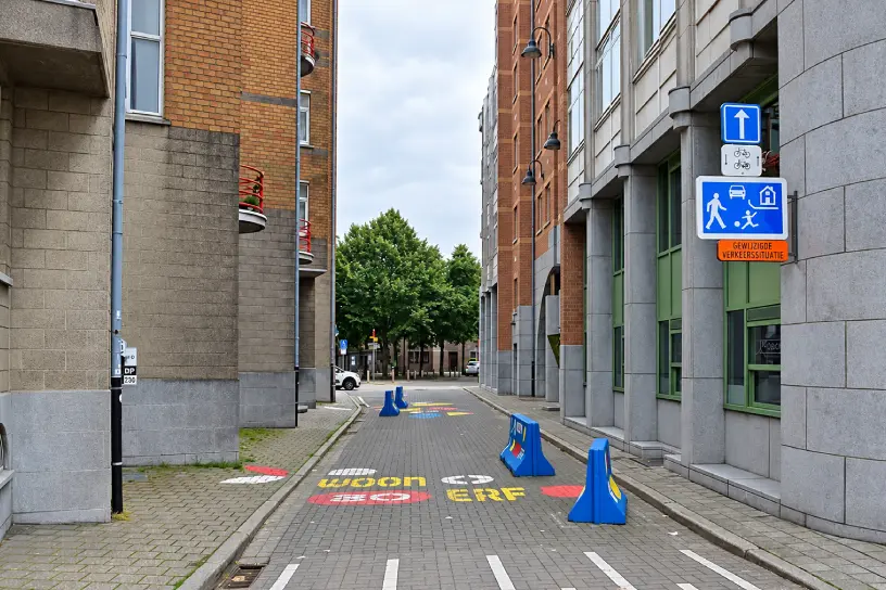 Rue étroite pavée à Anvers, avec un panneau bleu représentant un piéton, un enfant jouant au ballon, une voiture et une maison, sous une flèche blanche dirigée vers le haut.