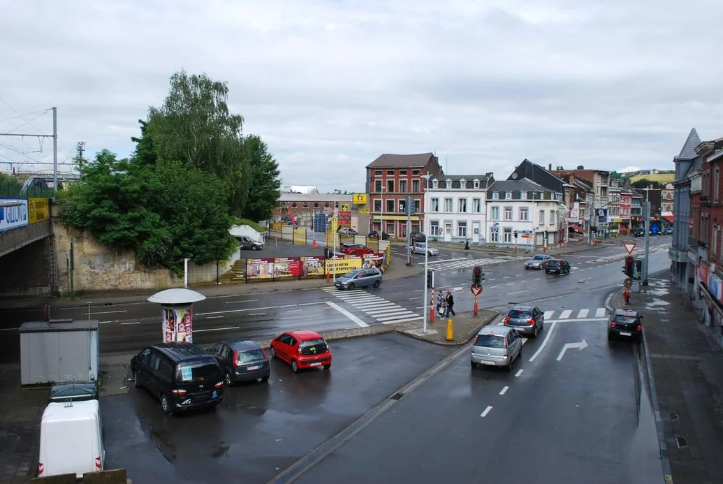 Carrefour à Chênée : voitures arrêtées aux feux rouges, signaux B1, flèche au sol vers la droite et passages piétons près d’un îlot où attendent des personnes.