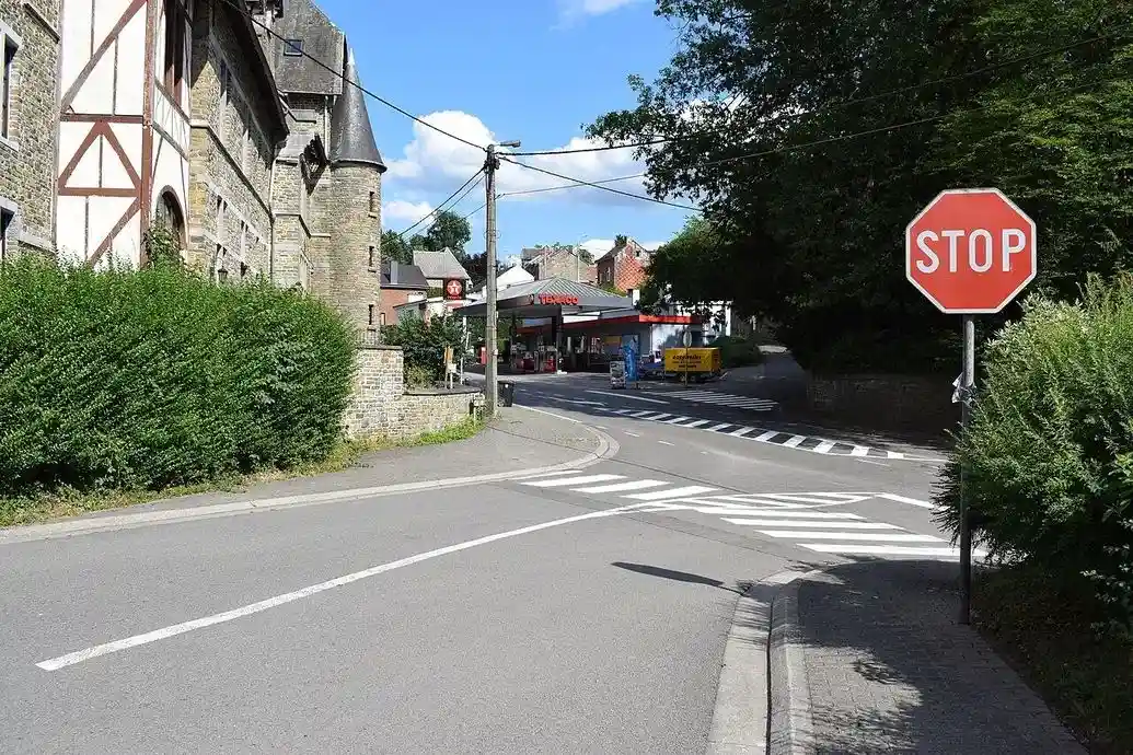 À Esneux, signal STOP à droite avant le carrefour, passage piéton sur la chaussée abordée et zone hachurée ; la végétation masque une partie de la vue à droite.