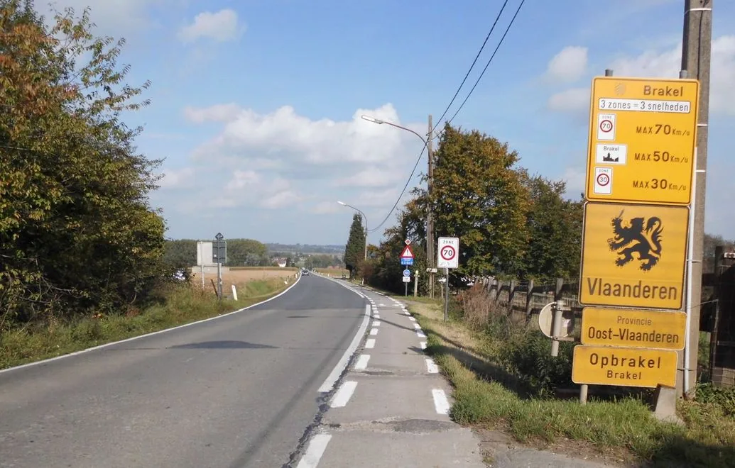 À Brakel, piste cyclable délimitée par deux lignes discontinues à droite ; panneau jaune récapitulatif, entrée ZONE 70, losange de priorité, signal A51 et D7.